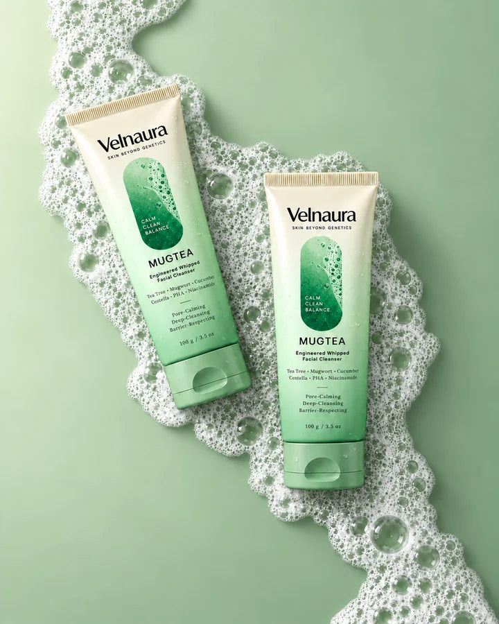 Two Velnaura Mugtea Engineered Whipped Facial Cleanser tubes, 100 g, lie side by side on a sage-green surface, resting on a winding ribbon of fine, close-knit white foam.