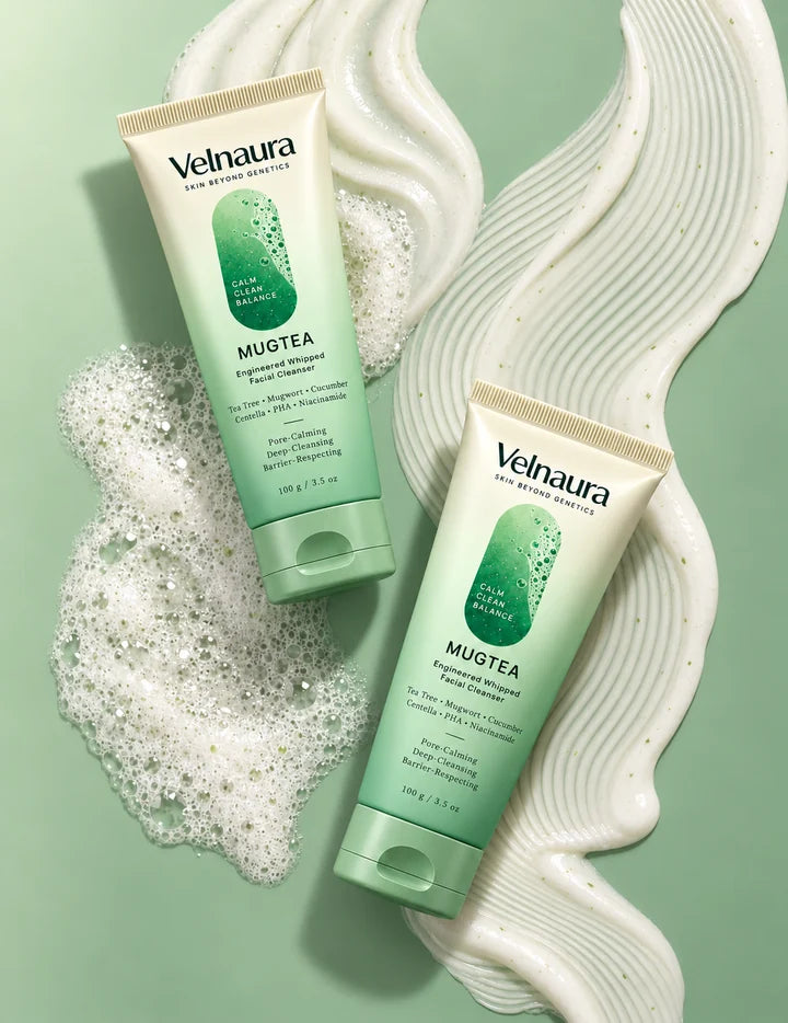 Two Velnaura Mugtea Engineered Whipped Facial Cleanser tubes lie diagonally on a soft sage-green surface, with white foam on the left and a wide, ridged ribbon of ivory cream flecked with tiny green specks between them.