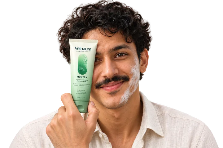 A smiling man with dark curly hair and a moustache holds a Velnaura Mugtea tube beside one eye, with a trail of soft white foam along his cheek.