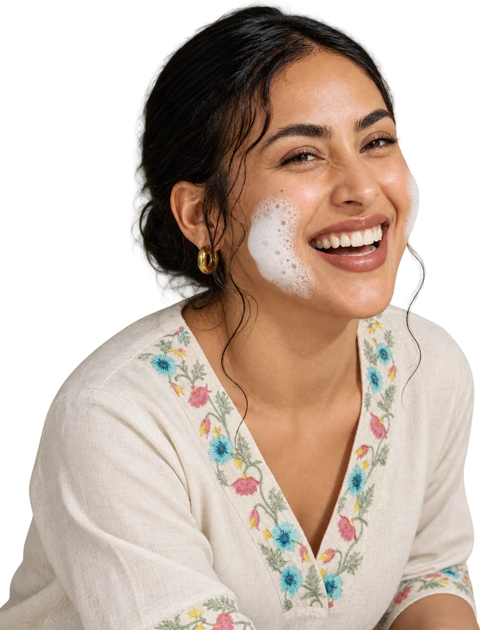 A woman with dark hair pinned back laughs with her head tilted, a patch of soft white foam on her cheek, wearing a cream linen top with floral embroidery at the neckline.