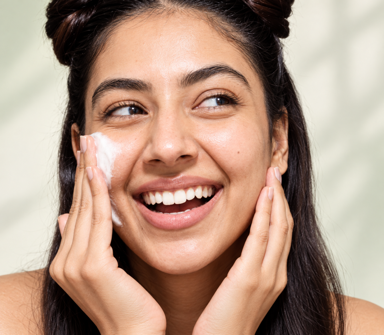 Smiling woman with dark hair in two buns, hands at her cheeks, with a little cleanser foam on one cheek.