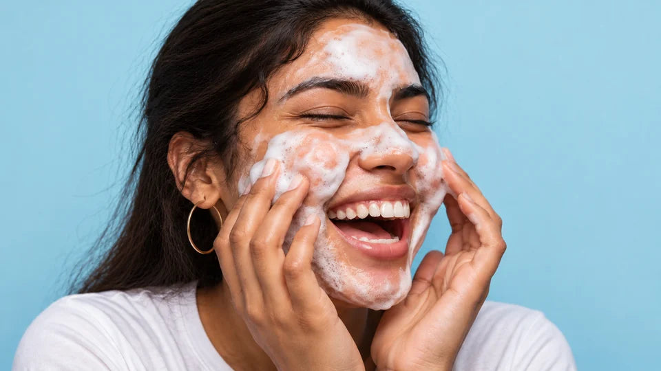 Laughing woman with straight dark hair massaging cleanser foam onto her cheeks against a blue background.