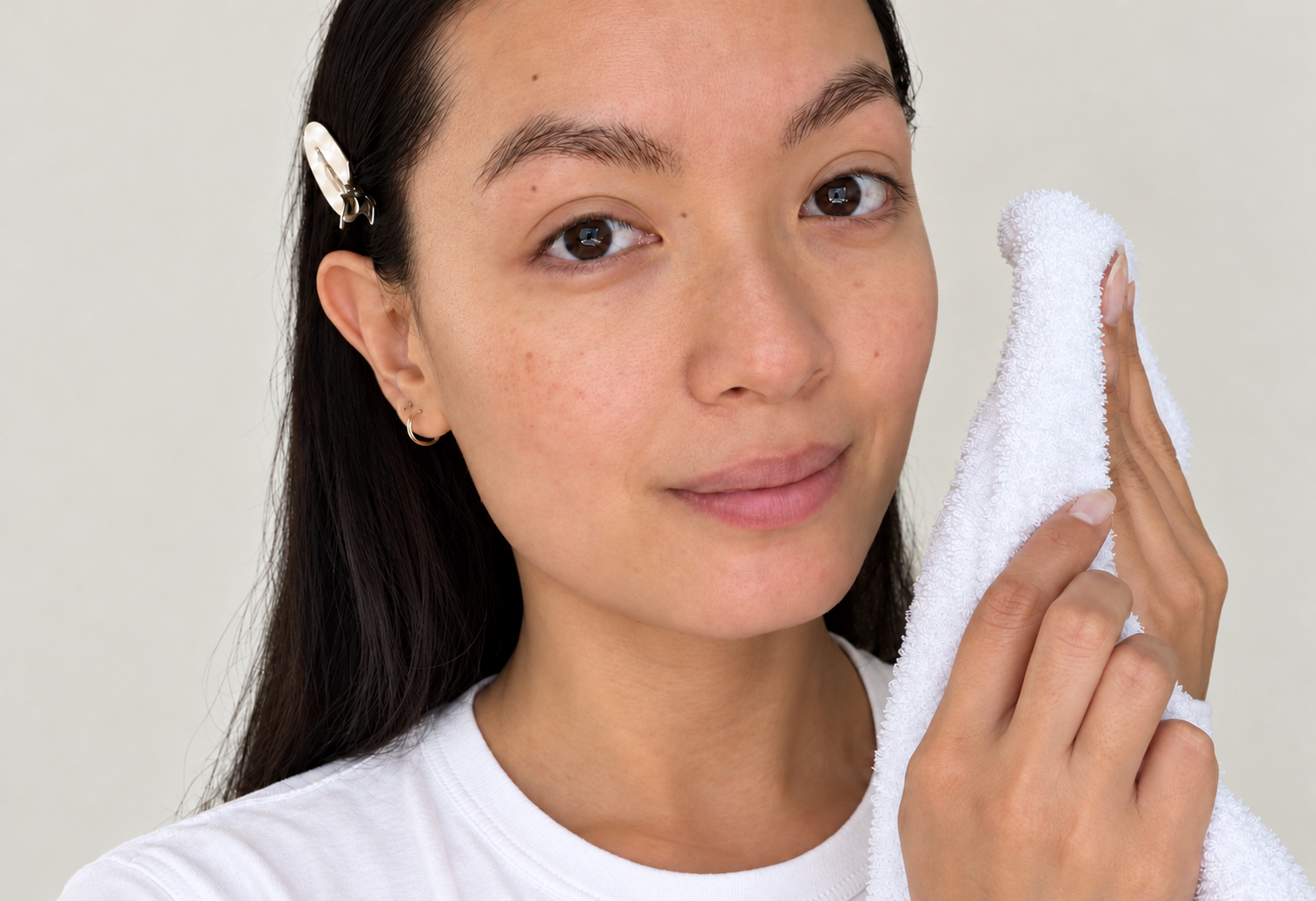 Woman with dark hair holding a white towel beside her cheek in soft natural window light.
