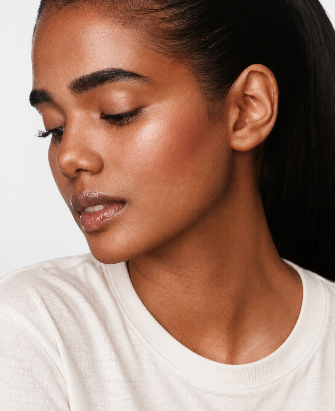 Close portrait of a woman with slicked-back dark hair looking down, wearing an off-white tee, with a natural glow on her cheek.